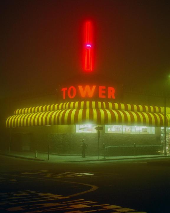 A person stands outside a store with a red and yellow striped awning and a neon sign that reads "Tower" in red.