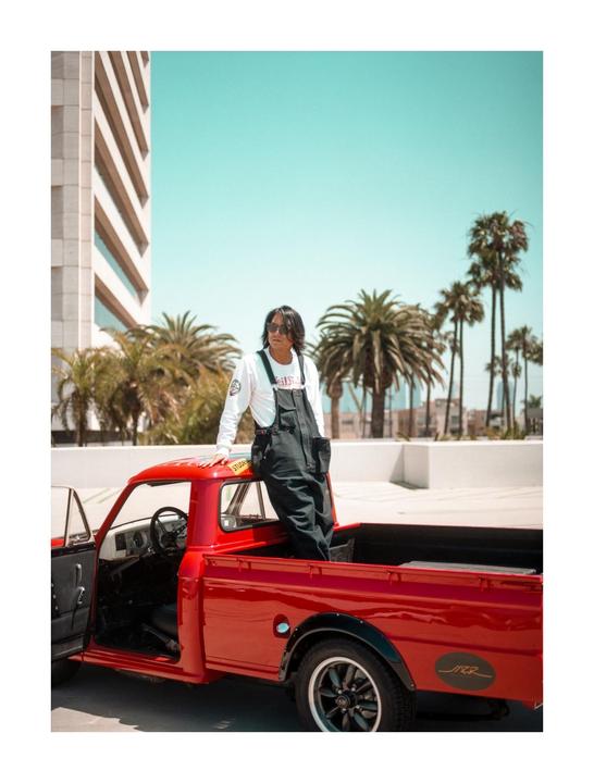 A man in overalls stands in the bed of a red pickup truck with palm trees and a building in the background.