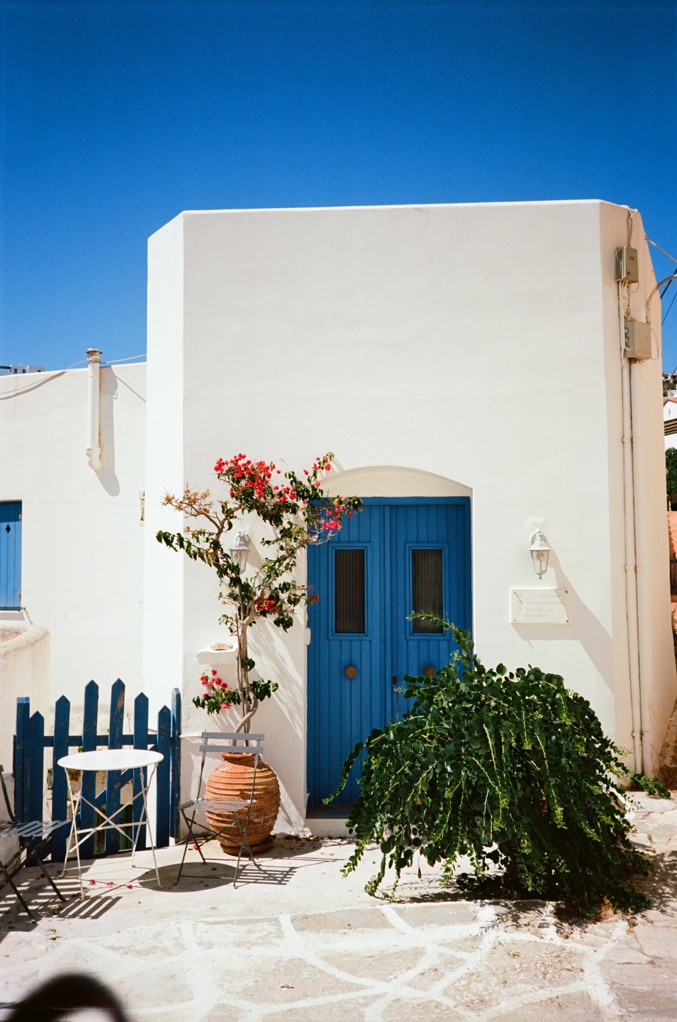 A white building with a blue door and a small patio with a table and chairs is visible under a clear blue sky.