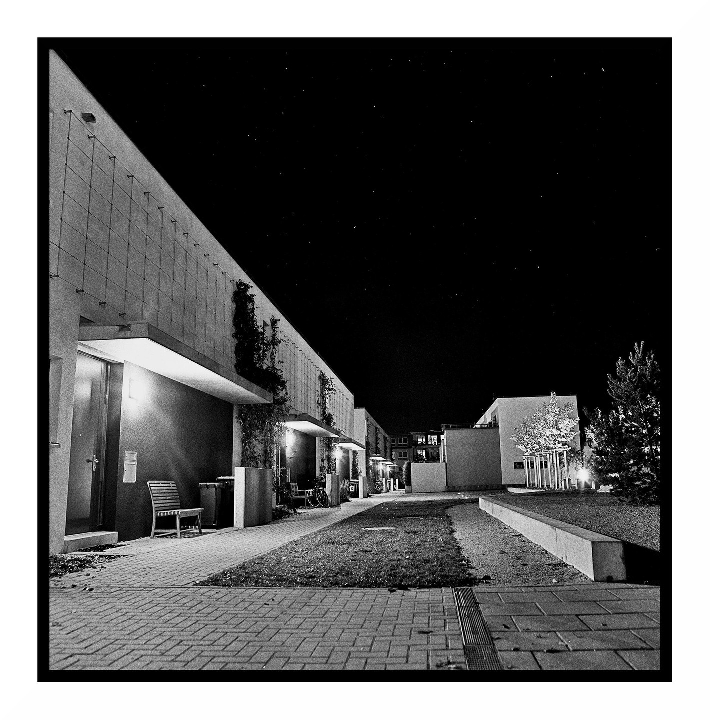A row of modern buildings with a paved walkway and grass is illuminated at night under a dark sky.