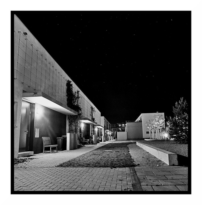 A row of modern buildings with a paved walkway and grass is illuminated at night under a dark sky.