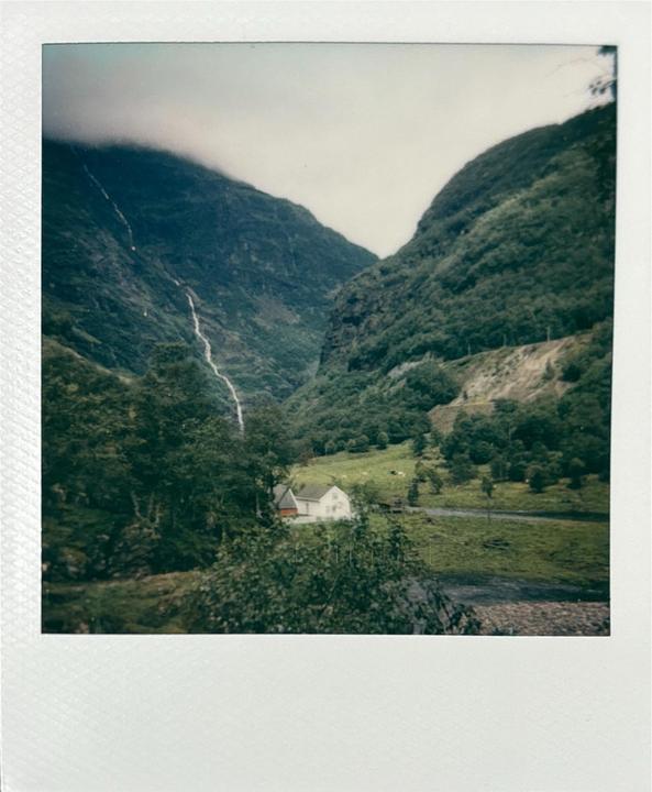 A small white house sits in a green valley with a waterfall cascading down a mountain in the background.