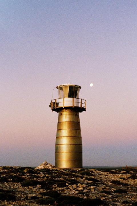 A golden lighthouse stands on a rocky landscape under a purple and blue sky with a visible moon.