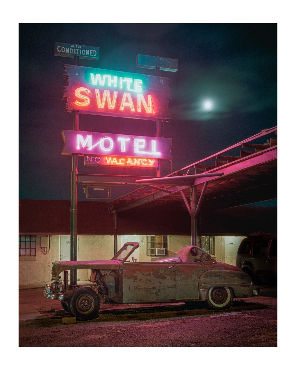 A neon sign for the White Swan Motel with a "No Vacancy" sign is illuminated at night next to a dilapidated car.