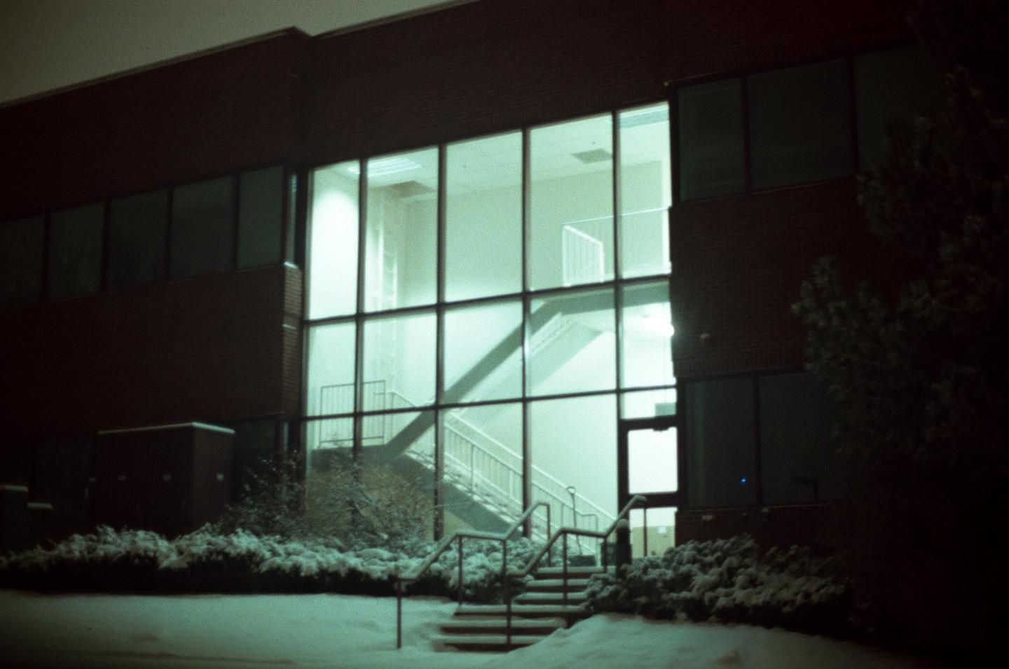A large glass window of a brick building reveals an interior staircase, with snow-covered ground and bushes in the foreground.