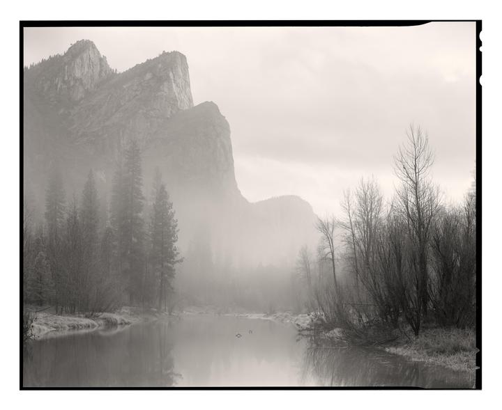 A misty mountain landscape features a river reflecting the sky and bare trees on its banks.