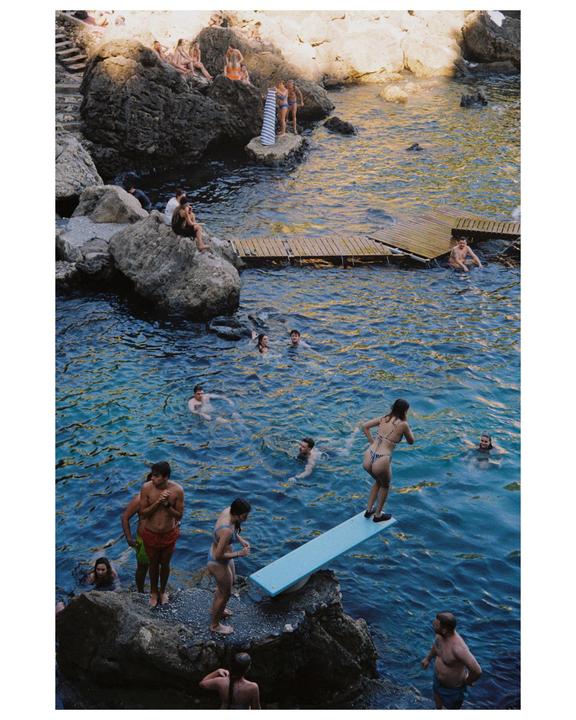 A woman stands on a diving board above a body of water with other people swimming and sitting on rocks.