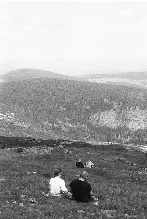 A couple sits in a grassy field overlooking a vast mountain landscape with forested hills.