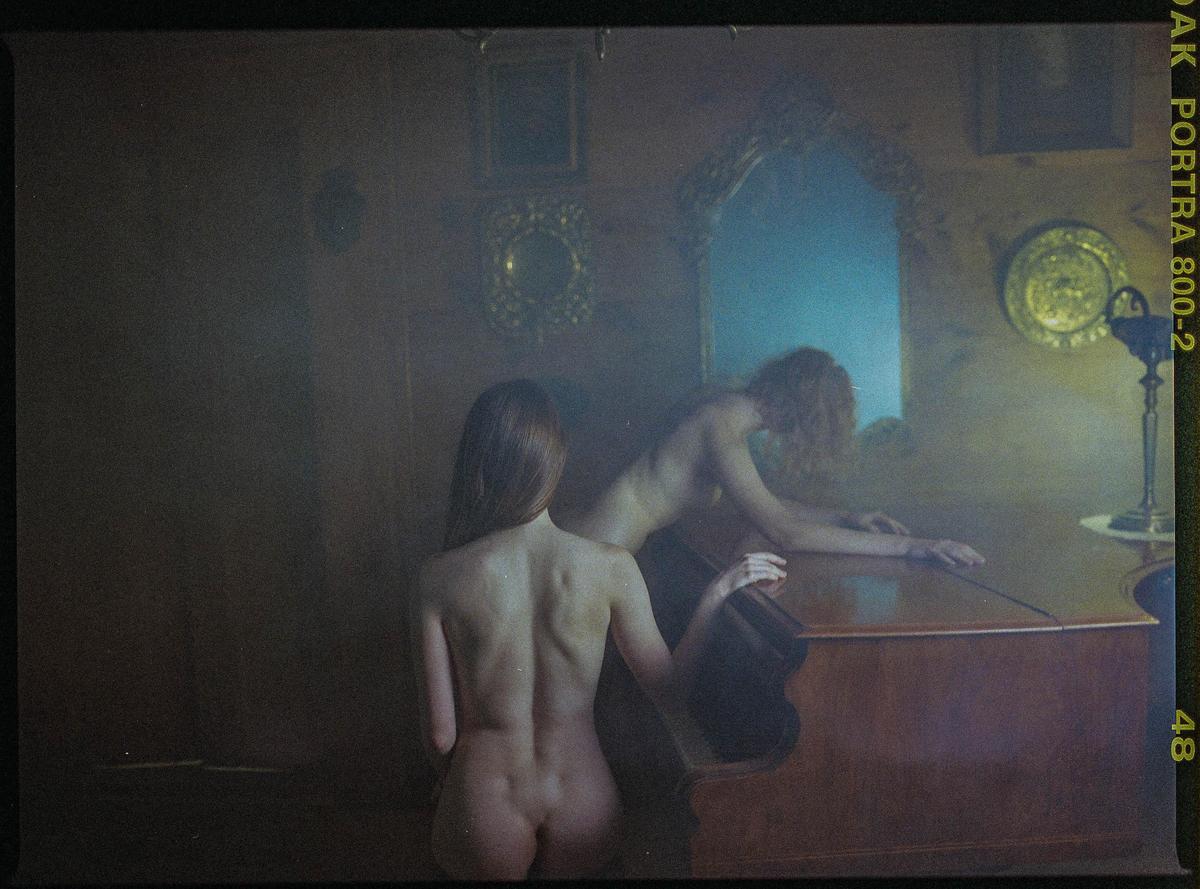 Two nude women are in a dimly lit room with one woman sitting in front of a piano and the other leaning over it.
