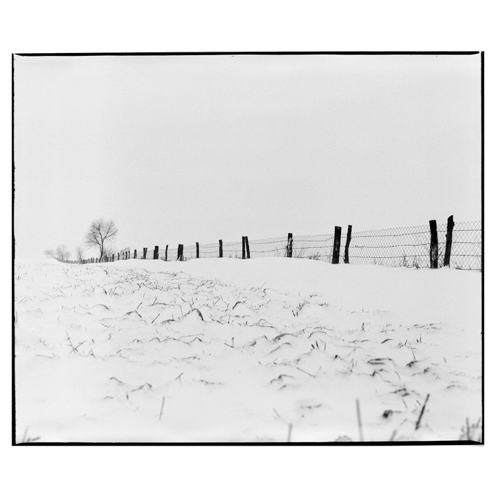 A wire fence with wooden posts stretches across a snow-covered field with bare trees in the distance.