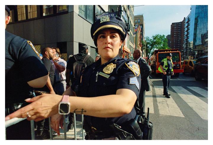 A woman police officer stands in the street with other people and vehicles in the background.
