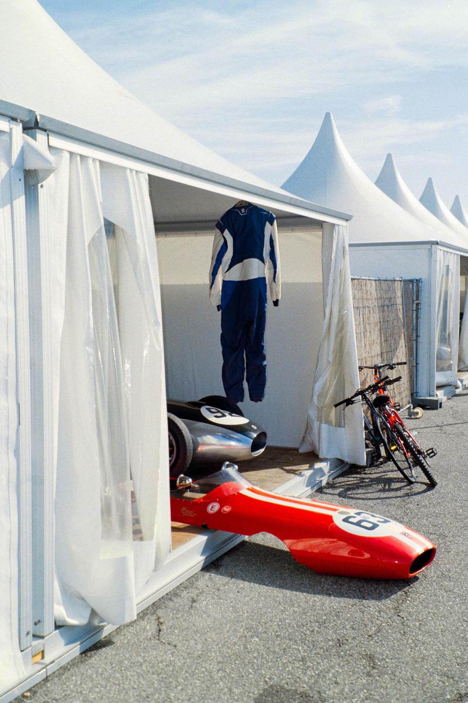 A racing suit hangs inside a white tent next to a red race car and a bicycle.