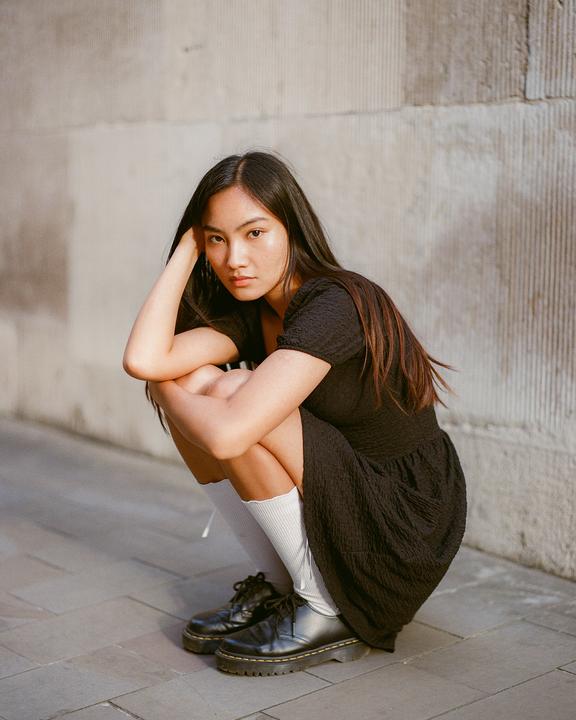 A woman in a black dress and white socks crouches on a paved surface next to a stone wall.