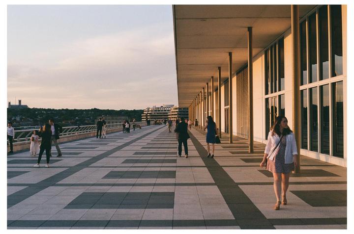 A woman walks along a long outdoor walkway next to a building with many windows, while other people gather in the distance.