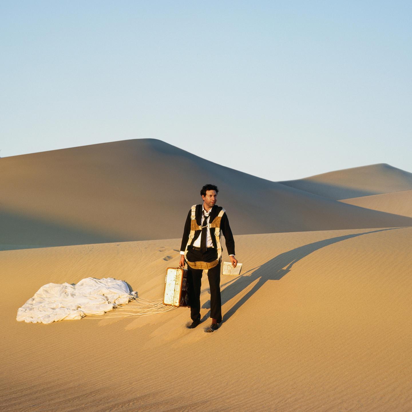 A man in a suit with a parachute harness stands in a desert landscape with sand dunes and a parachute on the ground.