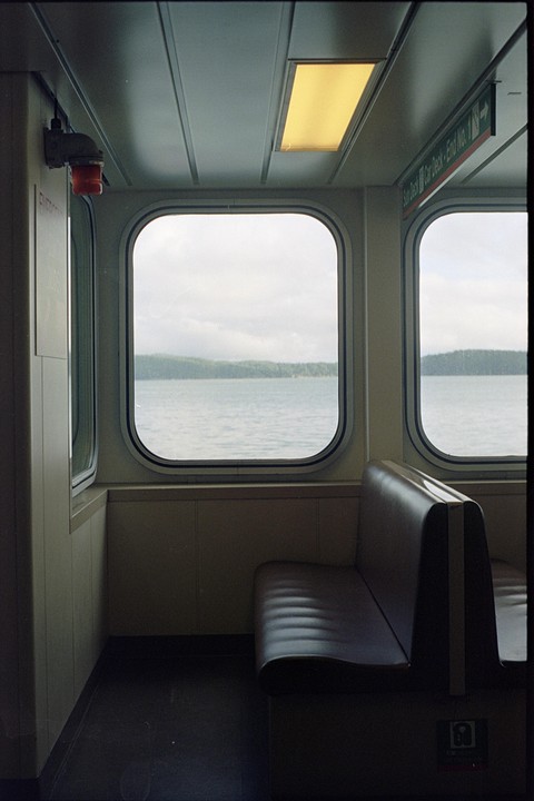 The interior of a ferry shows a bench seat and windows looking out onto a body of water with a distant tree-lined shore.
