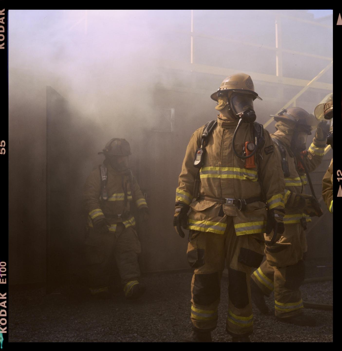 Three firefighters in full gear stand in a smoky room, with one in the foreground looking directly ahead.