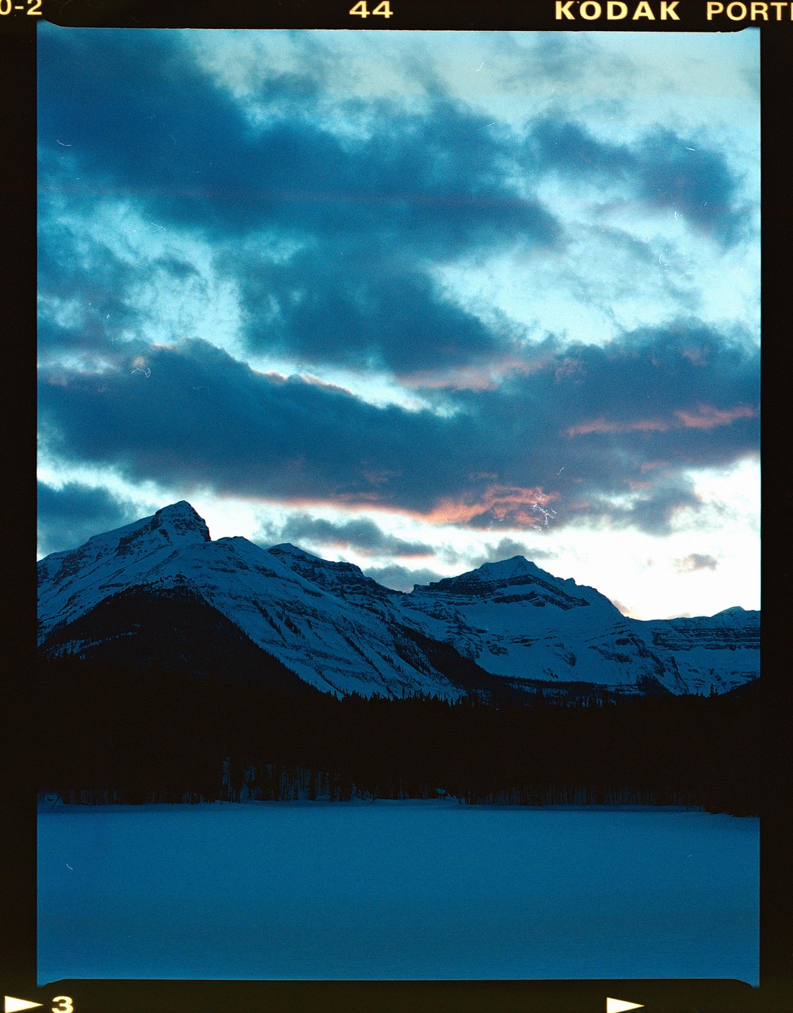 A snowy mountain range rises above a frozen lake under a cloudy sky with hints of pink.