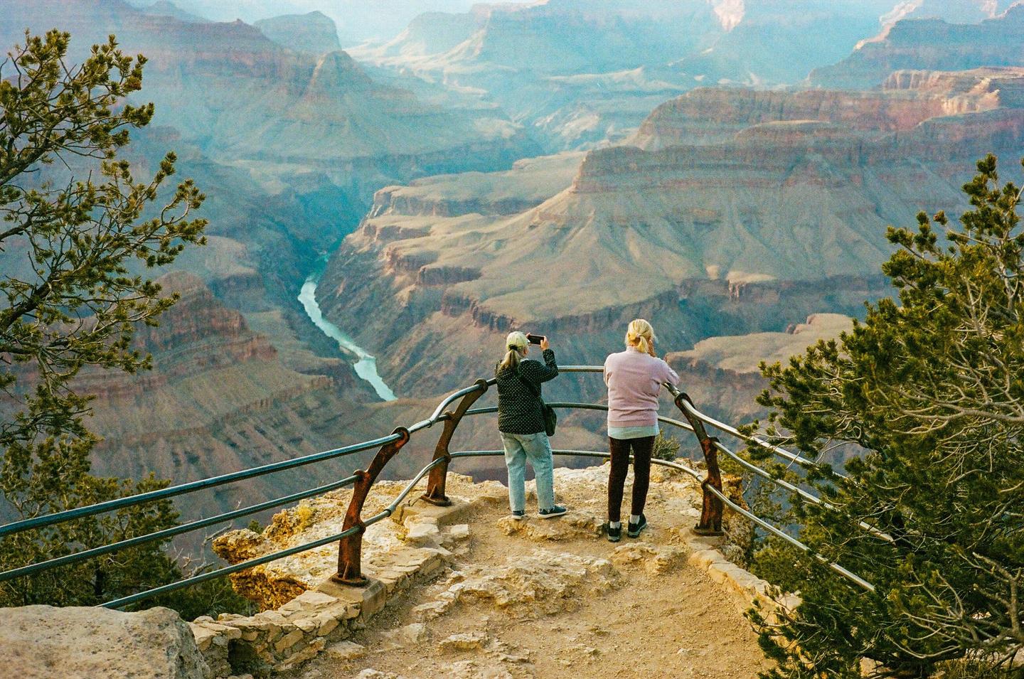 Two women stand at a railing overlooking a vast canyon with a river winding through it.