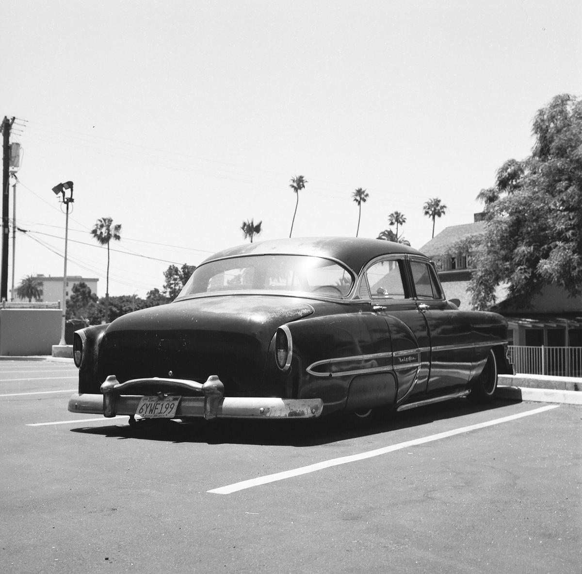 A black vintage car is parked in a parking lot with palm trees in the background.
