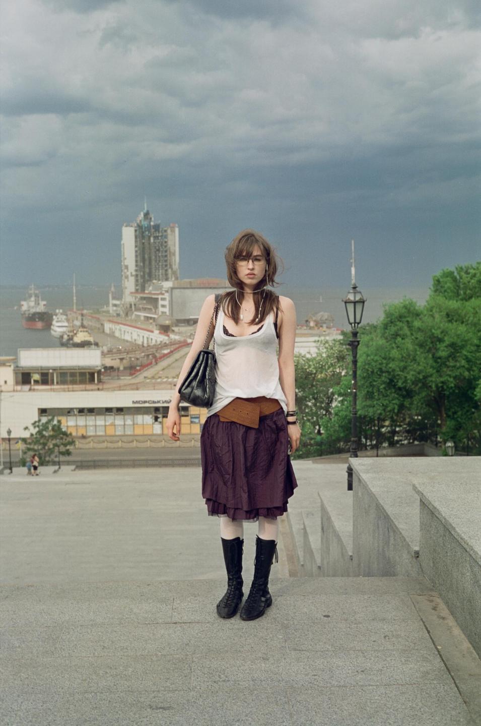 A woman stands on concrete steps overlooking a city harbor with a large building and ships under a cloudy sky.