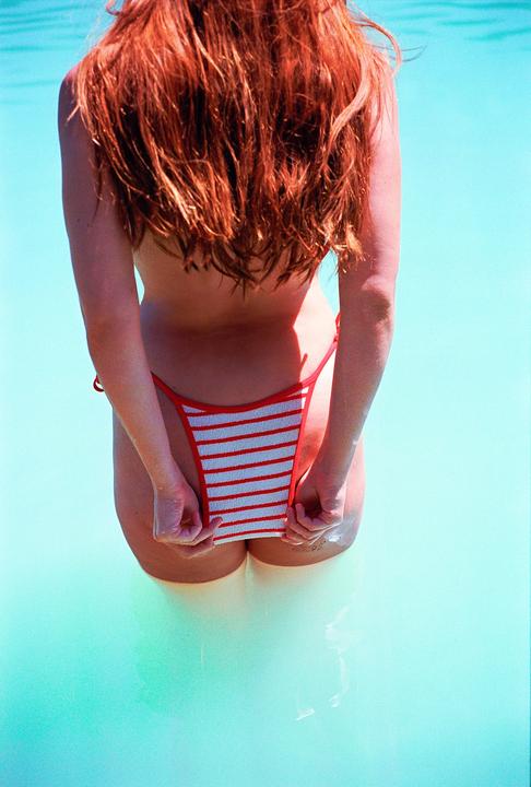 A woman with long red hair stands in turquoise water, pulling down her striped bikini bottom.