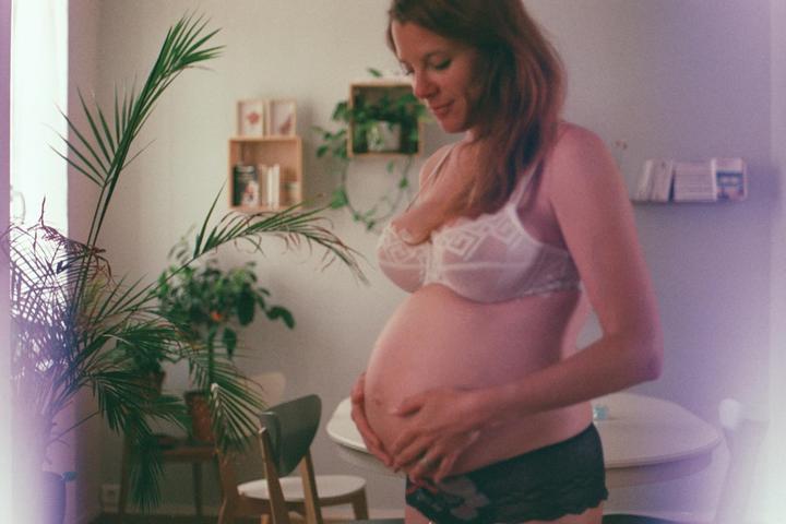 A pregnant woman in a white bra and black underwear holds her belly while standing in a room with plants.