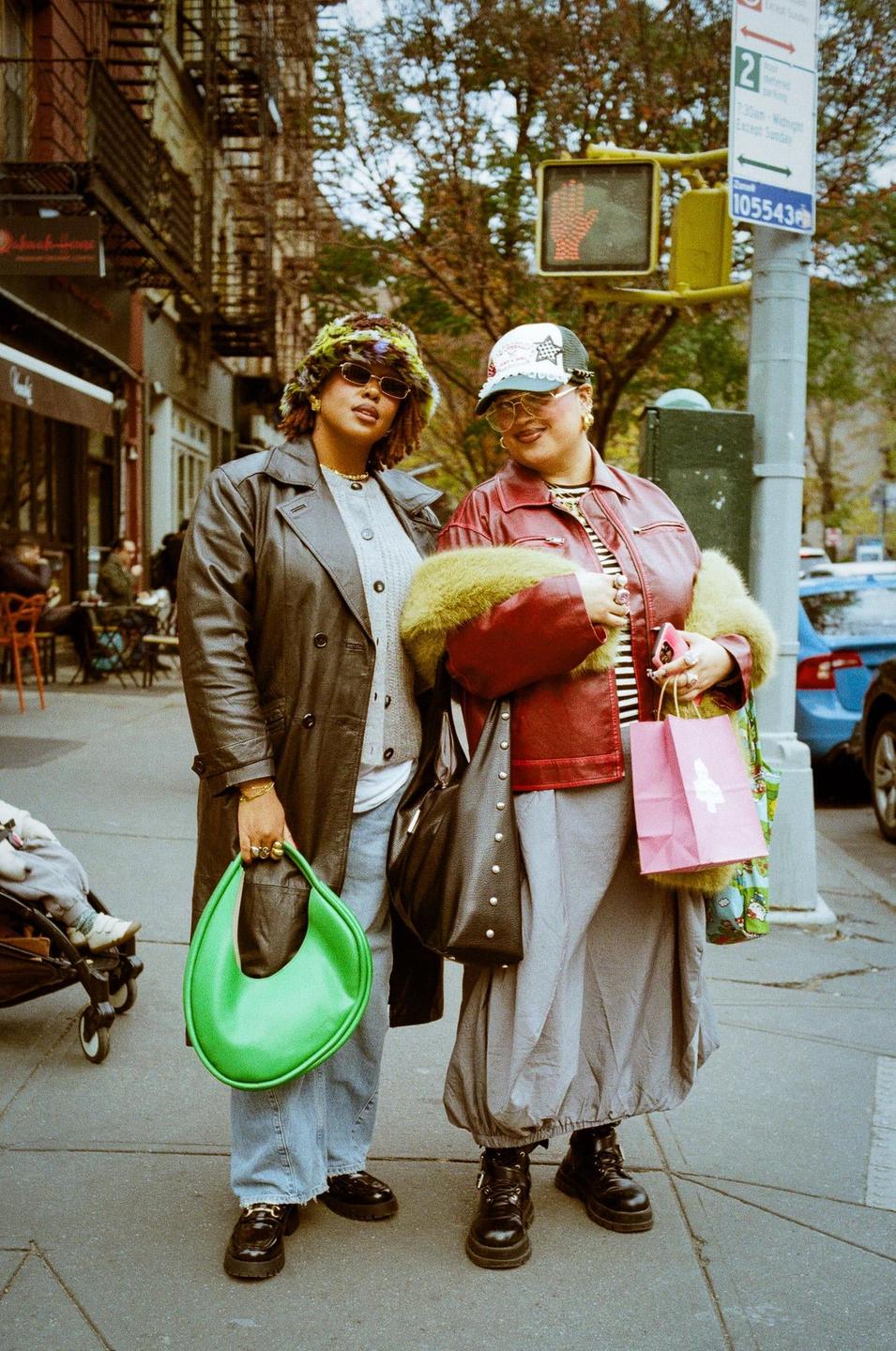 Two women stand on a city sidewalk, one holding a bright green handbag and the other a pink shopping bag.