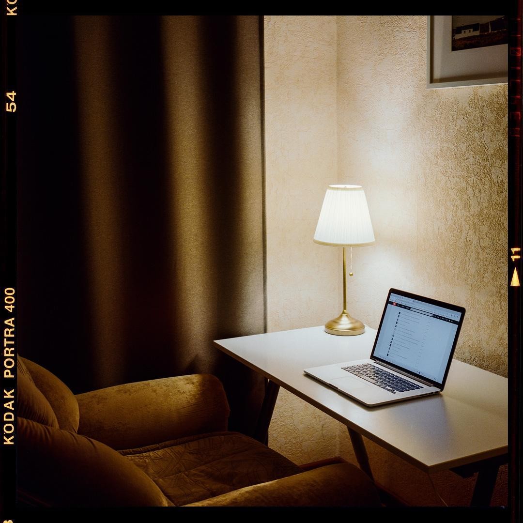 A laptop and a lamp sit on a white desk next to a brown armchair and curtain.