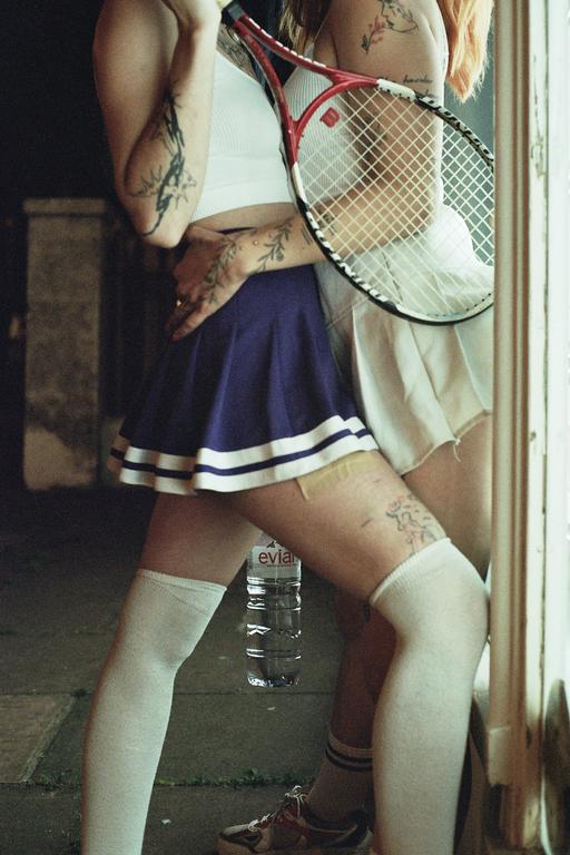 Two women in tennis skirts and knee-high socks stand close together, one holding a tennis racket.