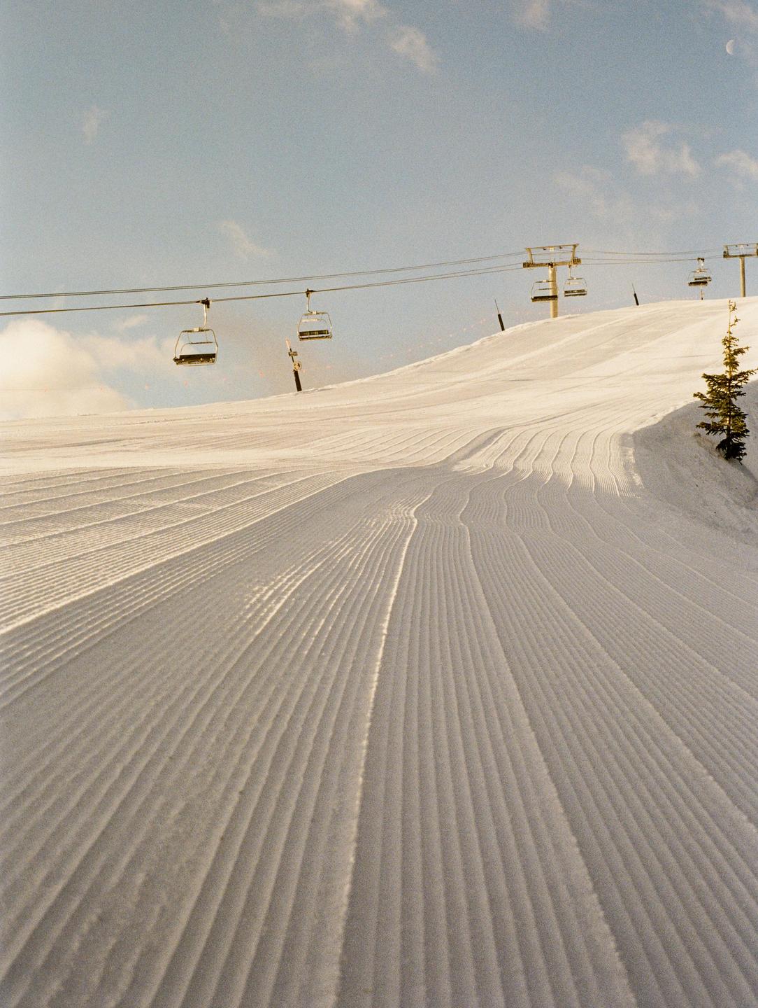 A ski slope with fresh corduroy snow is visible under a clear sky with ski lifts overhead.