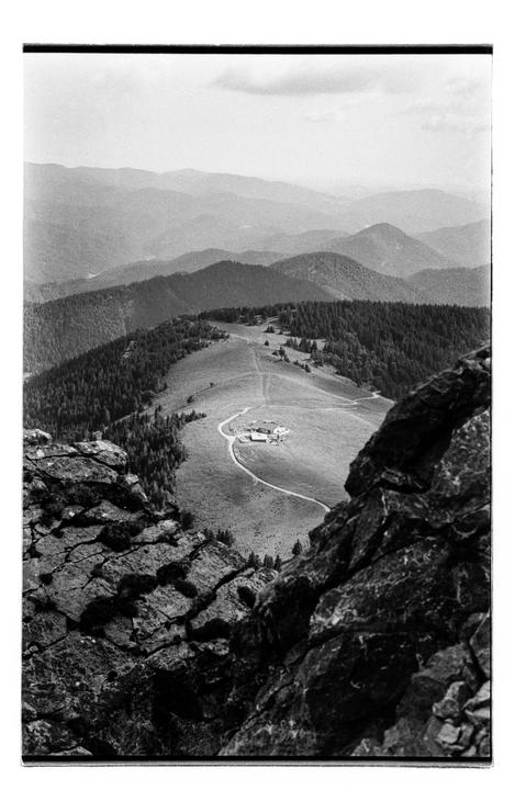 A house and winding road are nestled in a valley surrounded by mountains and forests, viewed from a rocky outcrop.