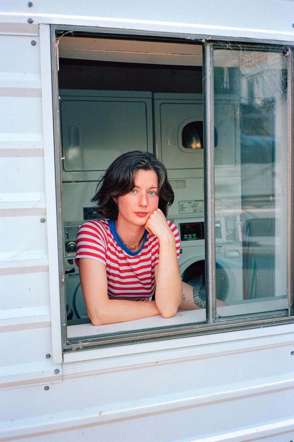 A woman in a striped shirt leans out of a window with washing machines visible in the background.