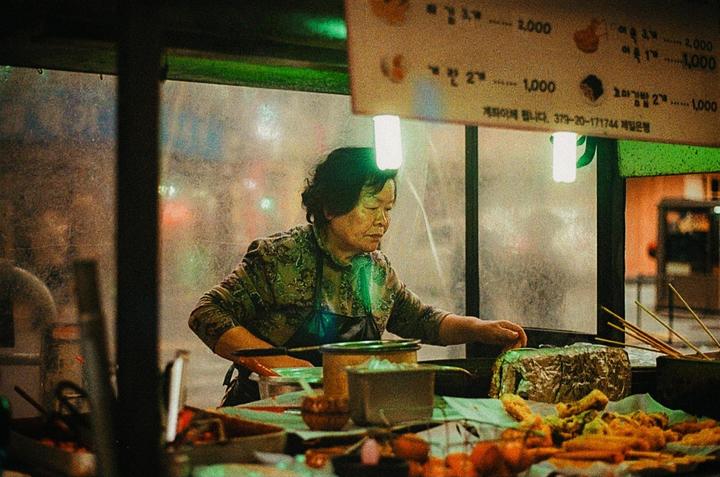 A woman prepares street food at night, illuminated by a bright light above her head.
