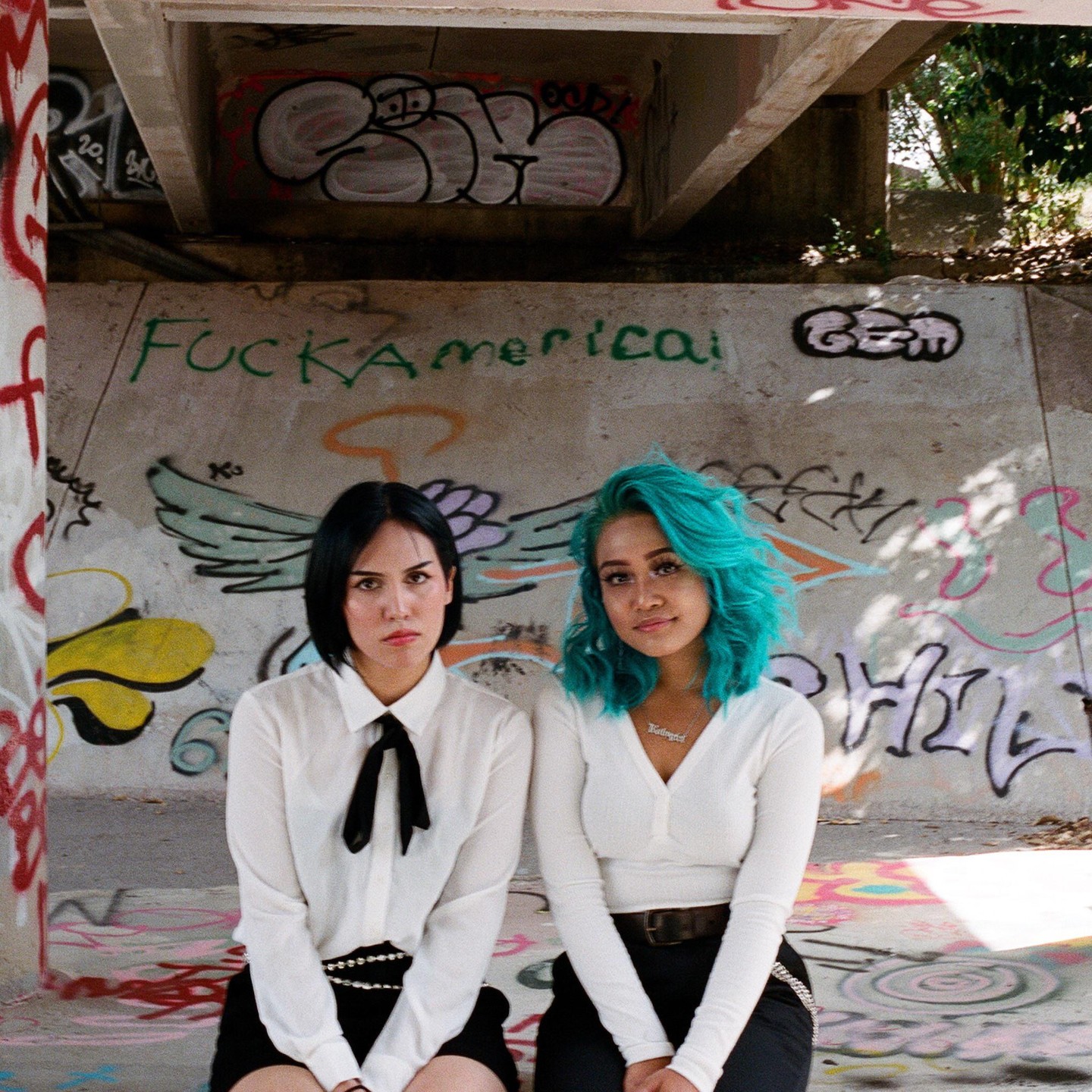 Two women sit in front of a graffiti-covered wall.