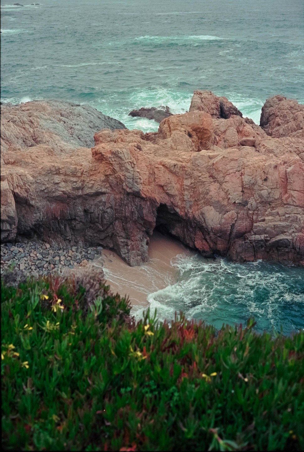 A hidden sandy cove with a sea cave is visible between rocky cliffs, with green foliage in the foreground.