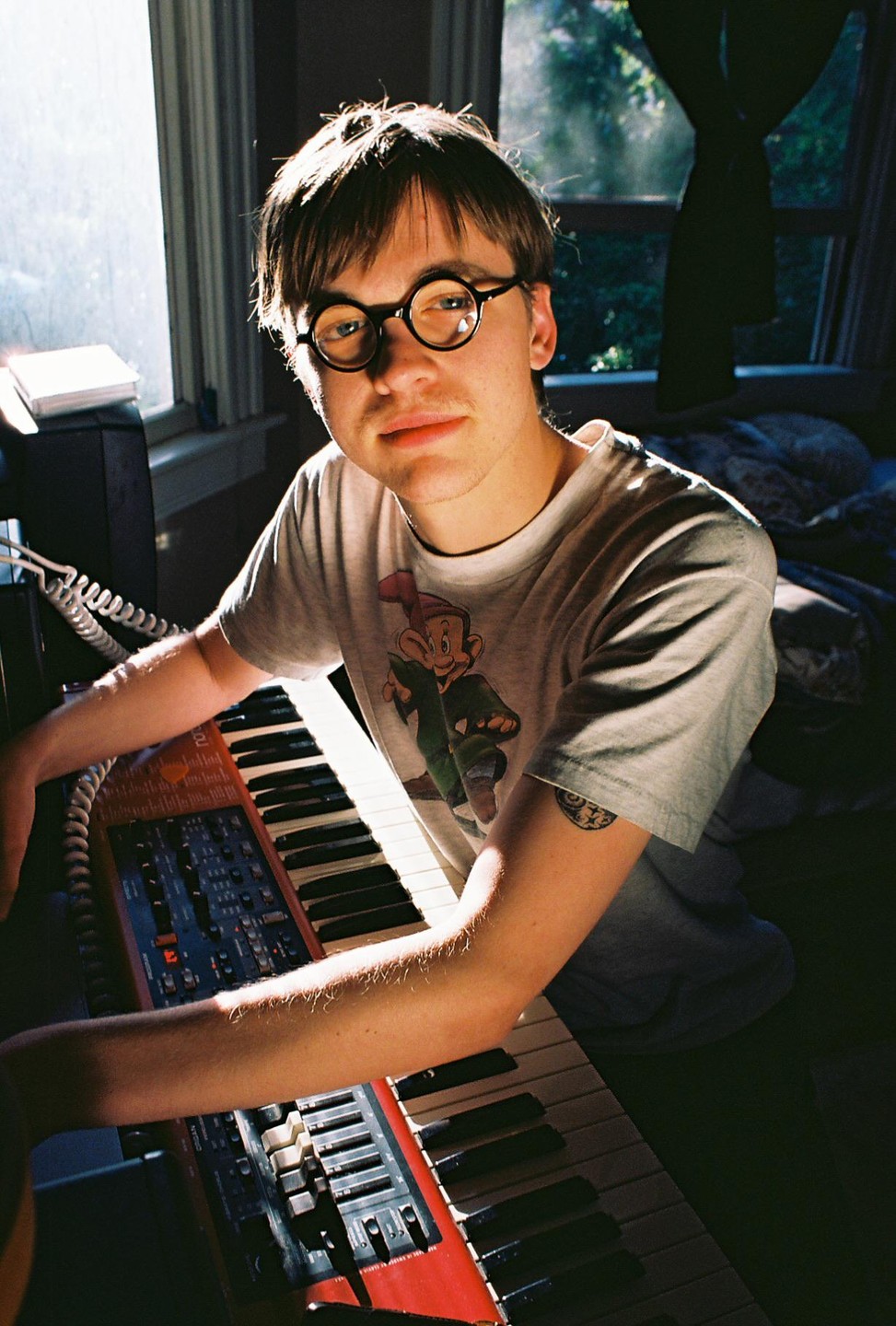 A young man with round glasses and a t-shirt with a cartoon character sits in front of a keyboard.