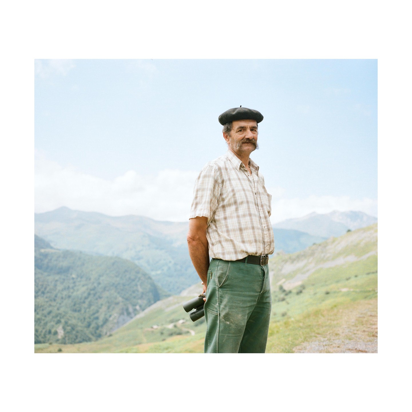 A man wearing a beret and plaid shirt stands on a mountain with binoculars behind his back.