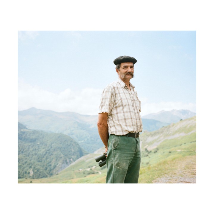 A man wearing a beret and plaid shirt stands on a mountain with binoculars behind his back.