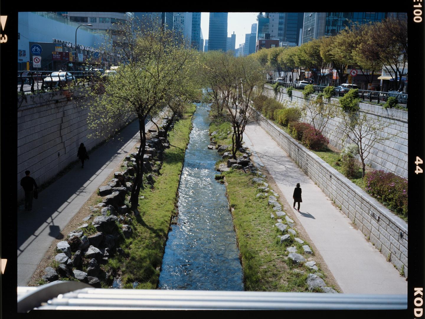 A narrow river flows between two paved walkways lined with trees and stone walls, with city buildings in the background.