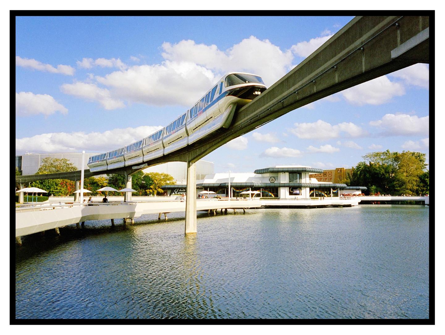 A monorail train travels on an elevated track over a body of water with buildings and trees in the background.