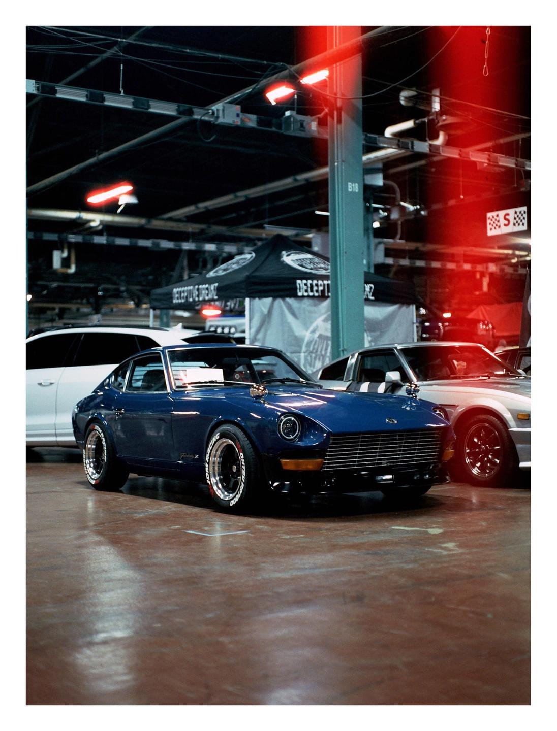 A blue Datsun 280Z is parked indoors with other vehicles and tents in the background.