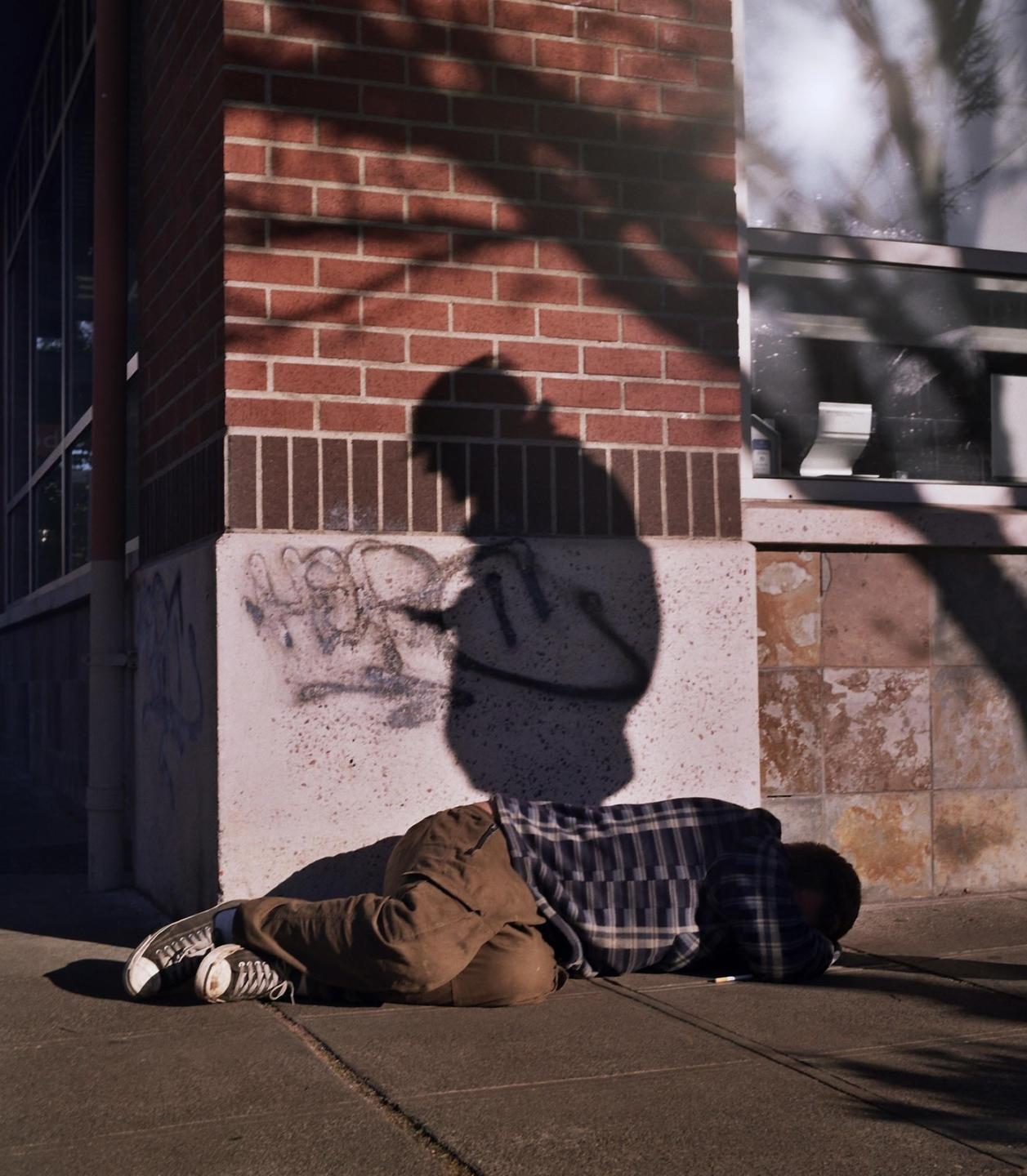 A person is lying on the sidewalk next to a brick building with graffiti and a large shadow of a person on the wall.