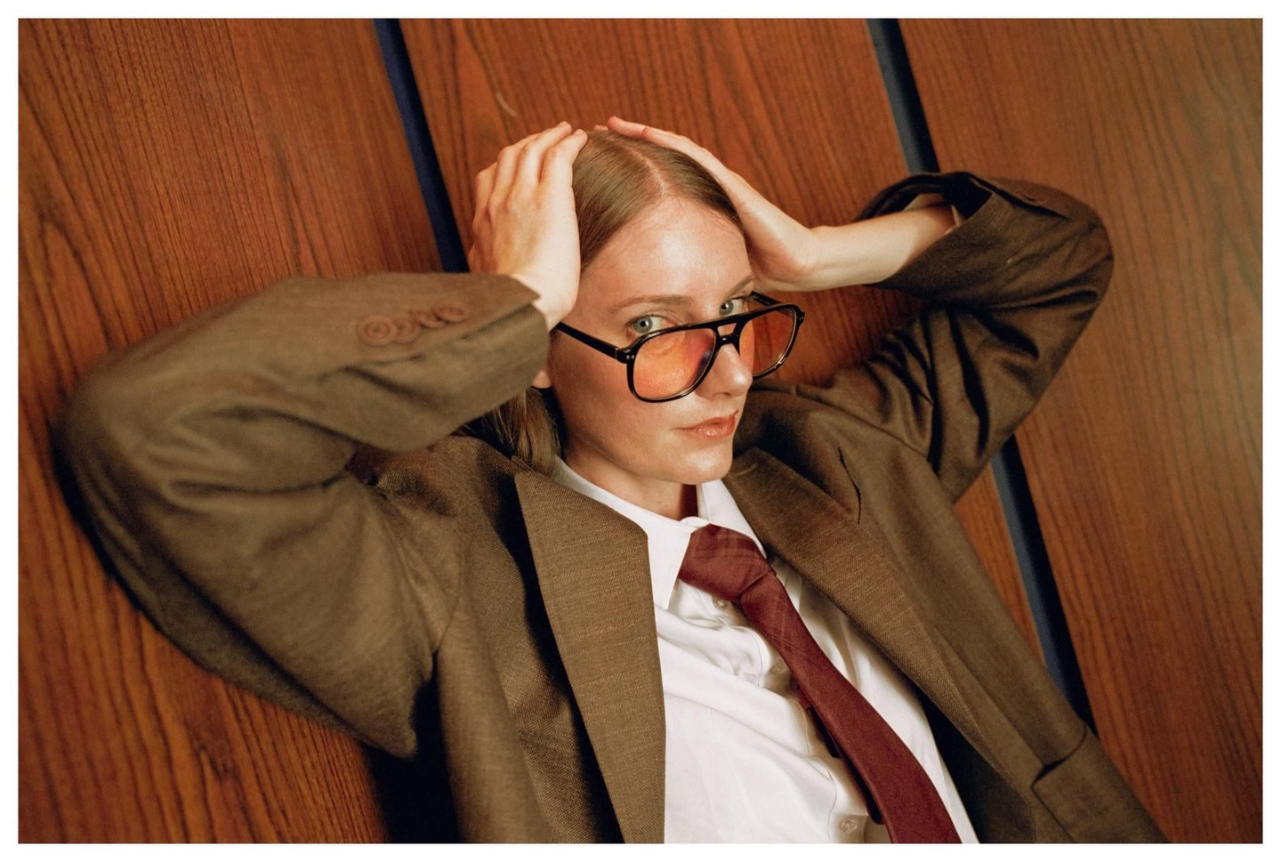 A woman wearing a suit, tie, and orange-tinted glasses lies on a wooden floor with her hands behind her head.