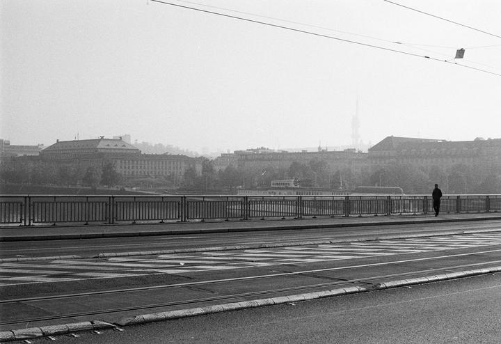A man walks on a bridge over a river with buildings and a hazy sky in the background.