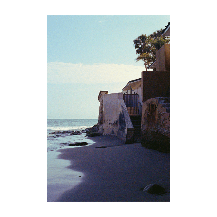A house with a gate and stairs sits on a sandy beach next to the ocean.