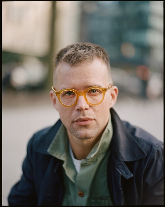 A man with short hair and yellow glasses looks directly at the viewer.