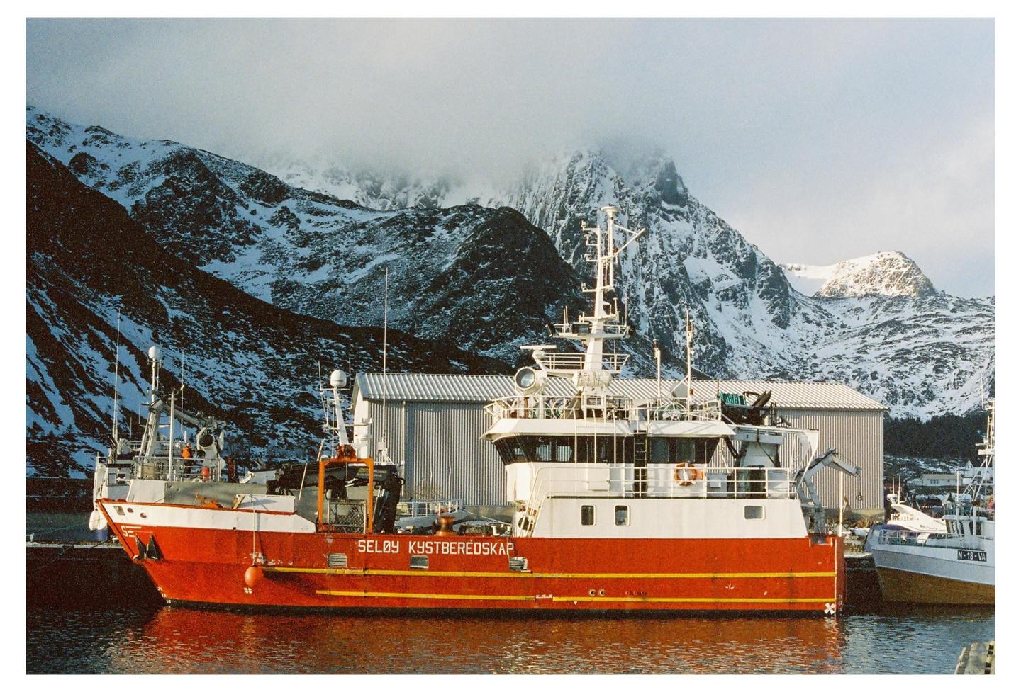 A red and white fishing boat is docked in a harbor with snow-covered mountains in the background.