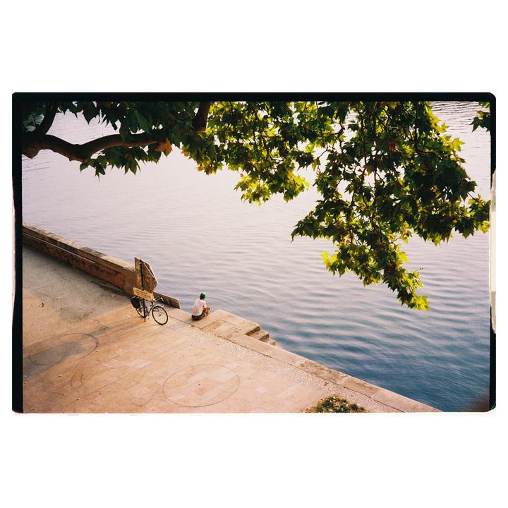 A man sits on the edge of a concrete embankment next to a body of water, with a bicycle parked nearby and a tree branch overhanging the scene.
