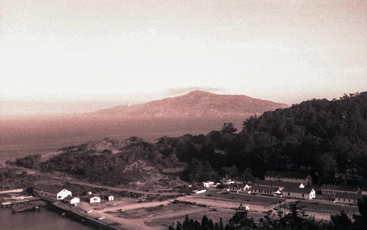A sepia-toned landscape shows a body of water with buildings along its shore and a large mountain in the background.